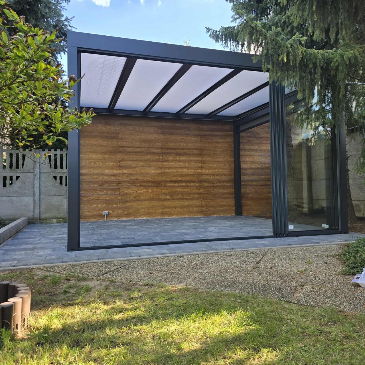 Pergola z kryciem stałym wyposażona w ramowy system szyb przesuwnych i rolety ekranowe. Zdjęcie jest własnością firmy Variant